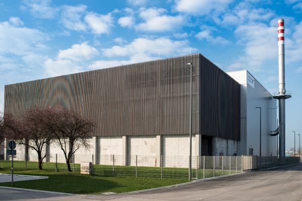 Das Holz-Heizkraftwerk in Heidelberg-Pfaffengrund können Interessierte bei einer Führung anlässlich der Energiewendetage am 15. September auch von innen kennen lernen. (Foto: Stadtwerke Heidelberg) Man sieht ein großes Gebäude mit einer kleinen Grünfläche davor. An dem Haus befindet sich ein Schornstein. (Foto: Stadtwerke Heidelberg)