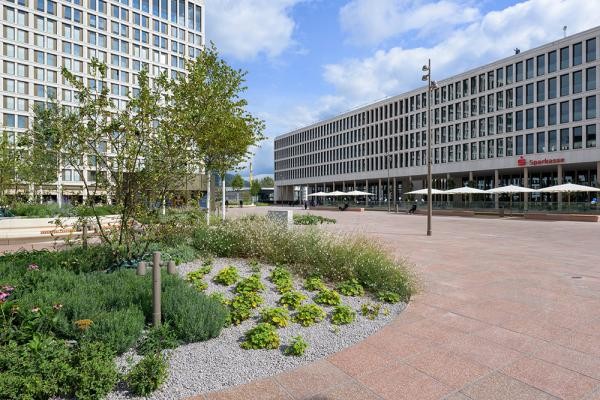 Ein moderner Platz mit modernen Gebäuden und grünen Inseln ist zu sehen 