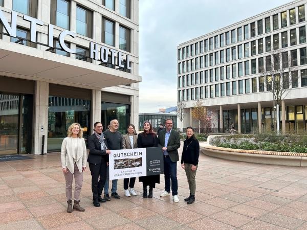 Vertreterinnen der Stadt Heidelberg und die Direktion des ALTANTIC Hotels übergaben die Preise an die Gewinnerinnen und den Gewinner (Mitte). (Foto: Stadt Heidelberg) Eine Gruppe von 7 Personen hält einen großen Gutschein und lacht in die Kamera. Im Hintergrund ein moderner Platz.