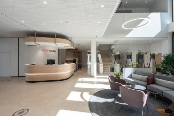 Rechts im Bild ist eine gemütliche Sitzecke mit Sofa und zwei Sesseln sowie einem grauen Teppich zu sehen. Dahinter stehen grüne Pflanzen. Links ist der helle Informationsschalter der Sparkassenfiliale zu sehen.