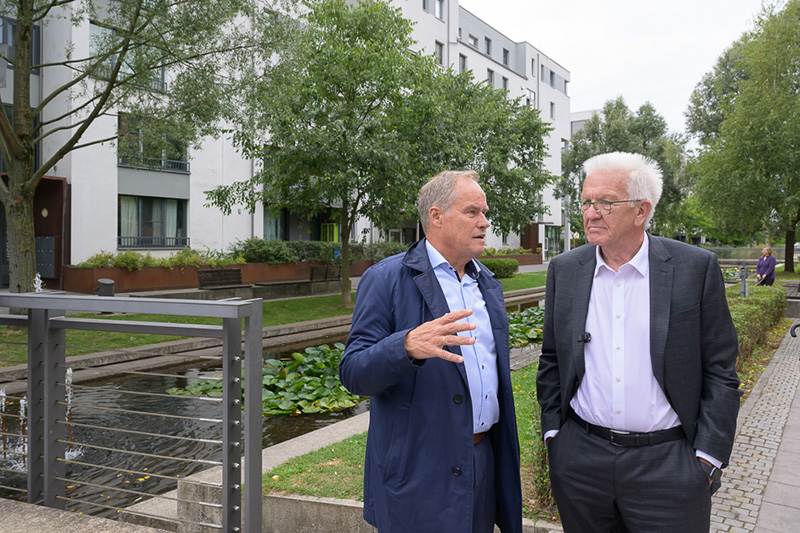 Zwei  Männer reden vor dem Hintergrund eines modernen Gebäudes und eines Parks miteinander.