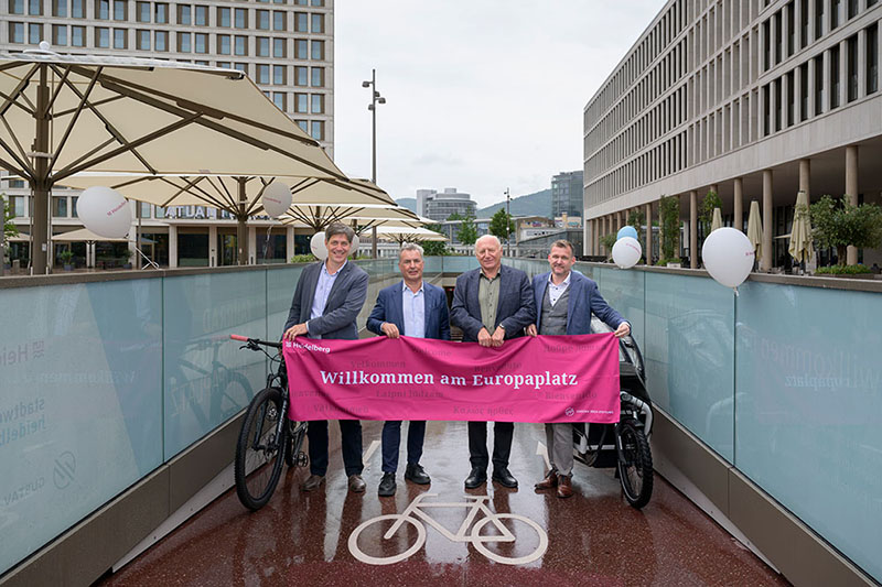 Vier Herren in Jackett posieren, im Eingang eines Fahrradparkhauses, mit einem pinken Banner mit der Aufschrift "Willkommen am Europaplatz."
